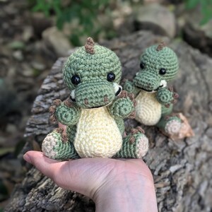 May include: Two crocheted green and cream dinosaur toys. The larger dinosaur is being held in a person's hand. The smaller dinosaur is sitting on a log.