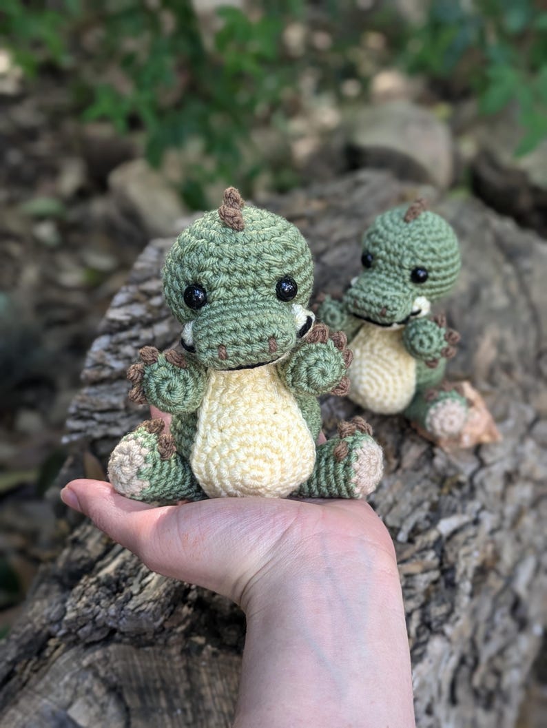 May include: Two crocheted green and cream dinosaur toys. The larger dinosaur is being held in a person's hand. The smaller dinosaur is sitting on a log.