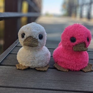 May include: Two plush duck toys, one white and one pink, with gold beaks and feet. The ducks are sitting on a wooden surface.