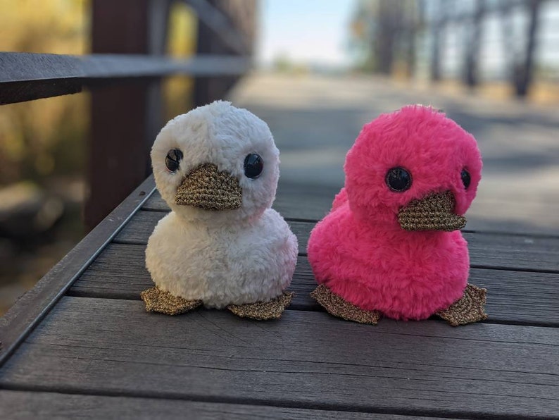 May include: Two plush duck toys, one white and one pink, with gold beaks and feet. The ducks are sitting on a wooden surface.