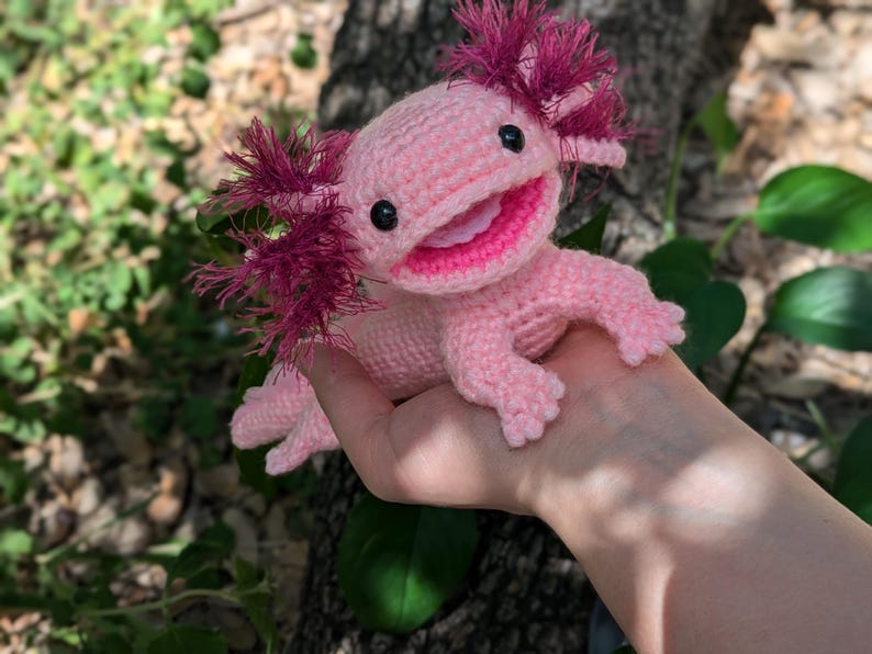 Może przedstawiać: R&oacute;żowa pluszowa zabawka axolotl wykonana na szydełku z czerwonymi, pierzastymi skrzelami. Zabawka ma szeroki uśmiech i czarne oczy.