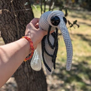 Peut inclure: Un jouet en peluche moustique gris et noir crocheté avec de grands yeux noirs. Le jouet est tenu par une main portant un bracelet rouge et jaune.