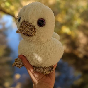 May include: A white plush duck with gold glitter accents and large, sparkly eyes. The duck is being held in a hand.