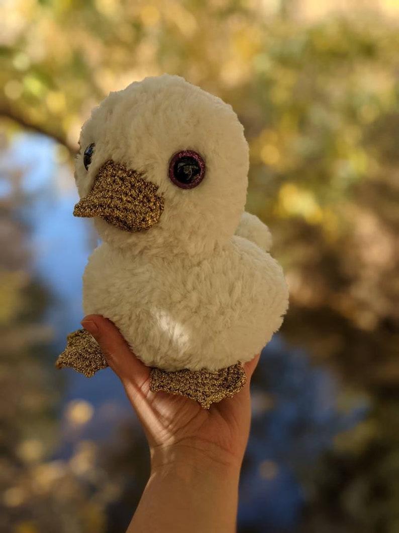 May include: A white plush duck with gold glitter accents and large, sparkly eyes. The duck is being held in a hand.