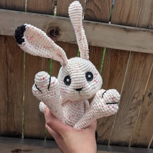 May include: A white crocheted bunny with pink speckles and black eyes. The bunny has long ears and is being held by a hand.