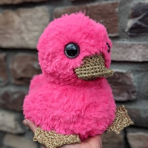 May include: A pink plush duck with gold glitter accents. The duck has large, black eyes and a fluffy body.