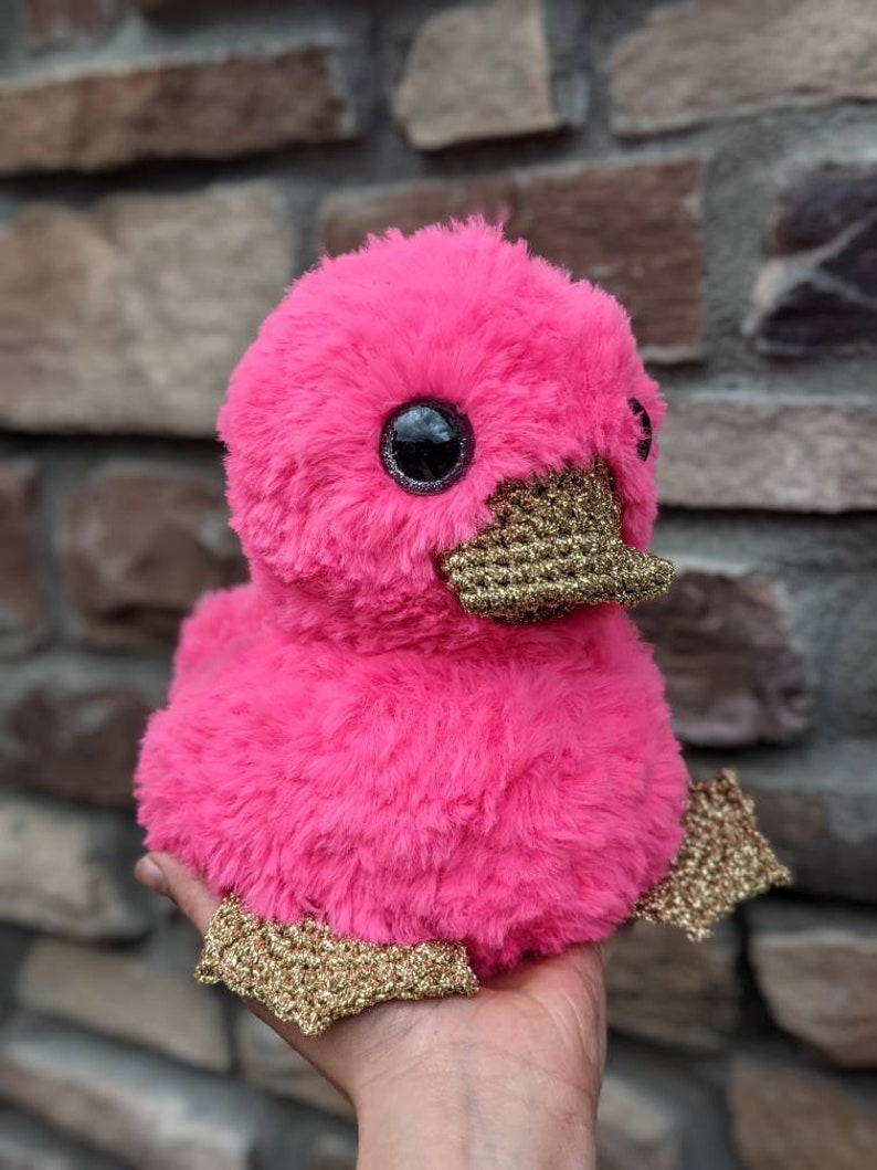 May include: A pink plush duck with gold glitter accents. The duck has large, black eyes and a fluffy body.