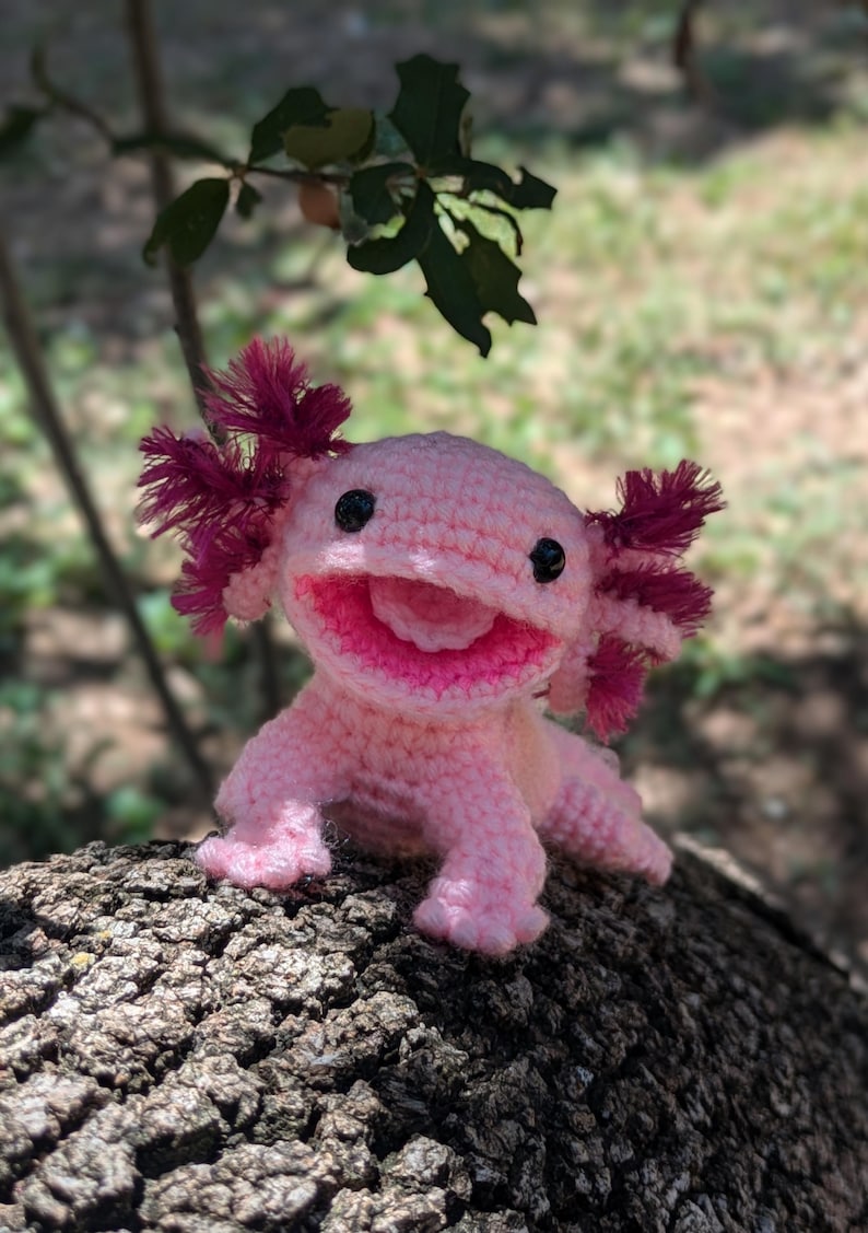 Może przedstawiać: R&oacute;żowa pluszowa zabawka axolotl wykonana na szydełku z czerwonymi skrzelami, czarnymi oczami i szerokim, zębatym uśmiechem. Zabawka siedzi na pniu drzewa.