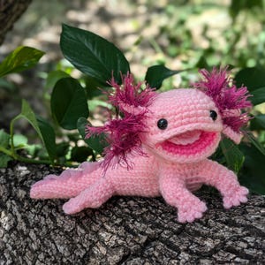 May include: A pink crocheted axolotl plush toy with pink and white details. The toy has a wide smile and large, black eyes. The axolotl is sitting on a tree branch.