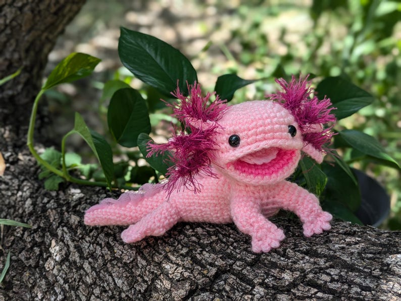 Może przedstawiać: R&oacute;żowa pluszowa zabawka axolotl wykonana na szydełku z r&oacute;żowymi i białymi detalami. Zabawka ma szeroki uśmiech i duże, czarne oczy. Axolotl siedzi na gałęzi drzewa.