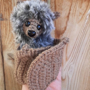 Pode incluir: Um pequeno morcego de crochê cinza e marrom com olhos pretos. O morcego está sentado em uma bolsa de crochê marrom.