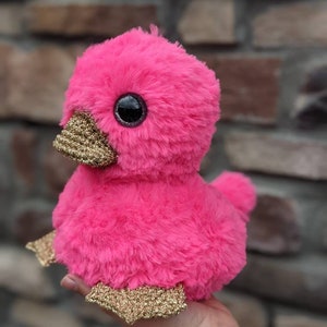 May include: A pink plush duck with gold glitter accents on its beak and feet. The duck has large black eyes and a fluffy pink body.