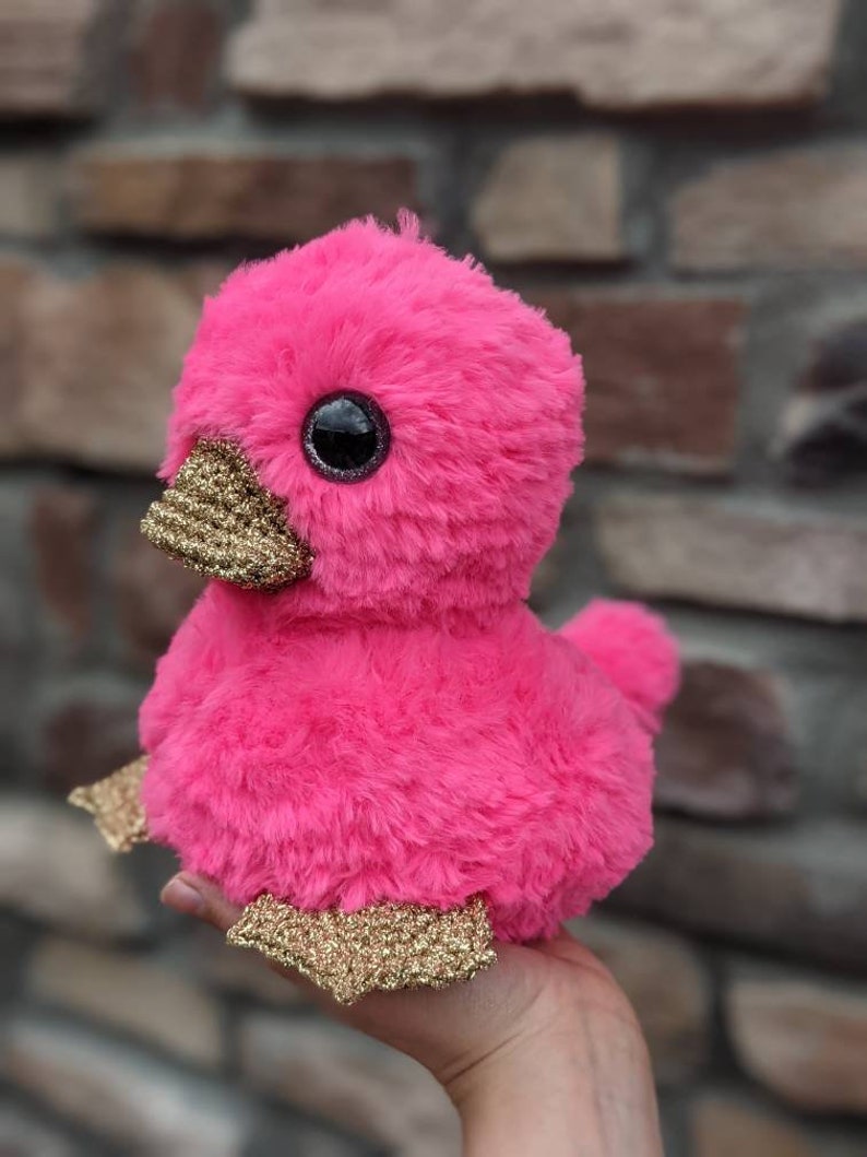 May include: A pink plush duck with gold glitter accents on its beak and feet. The duck has large black eyes and a fluffy pink body.