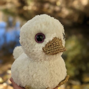 May include: A white plush duck with gold accents. The duck has large pink eyes and a gold beak. The duck is sitting on a hand.