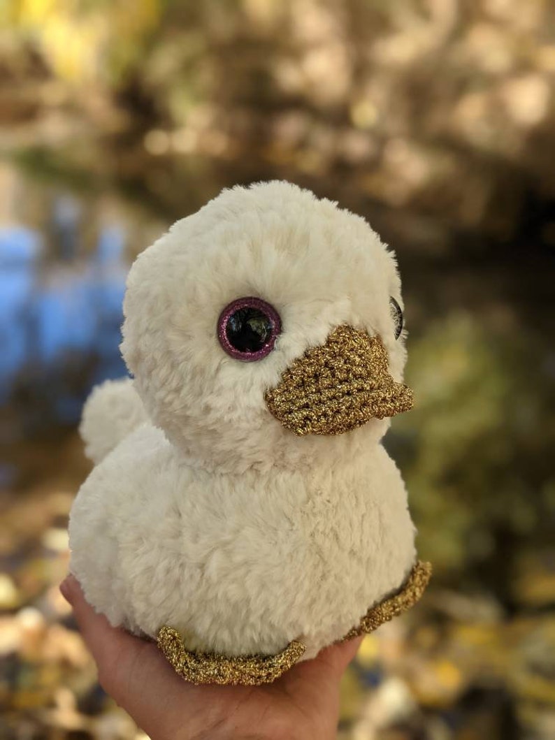 May include: A white plush duck with gold accents. The duck has large pink eyes and a gold beak. The duck is sitting on a hand.