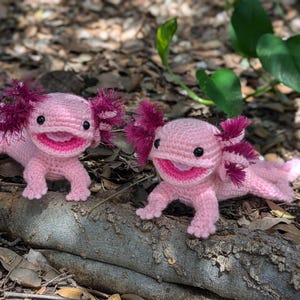 Może przedstawiać: Dwa r&oacute;żowe pluszowe aksolotlki wykonane na szydełku z r&oacute;żowymi i fioletowymi falbankami na głowach. Axolotki uśmiechają się i mają czarne oczy.
