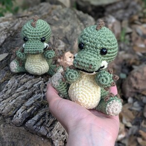 May include: Two crocheted green dinosaur plushies with brown accents. The smaller dinosaur is sitting on a tree stump, while the larger dinosaur is being held in a hand.