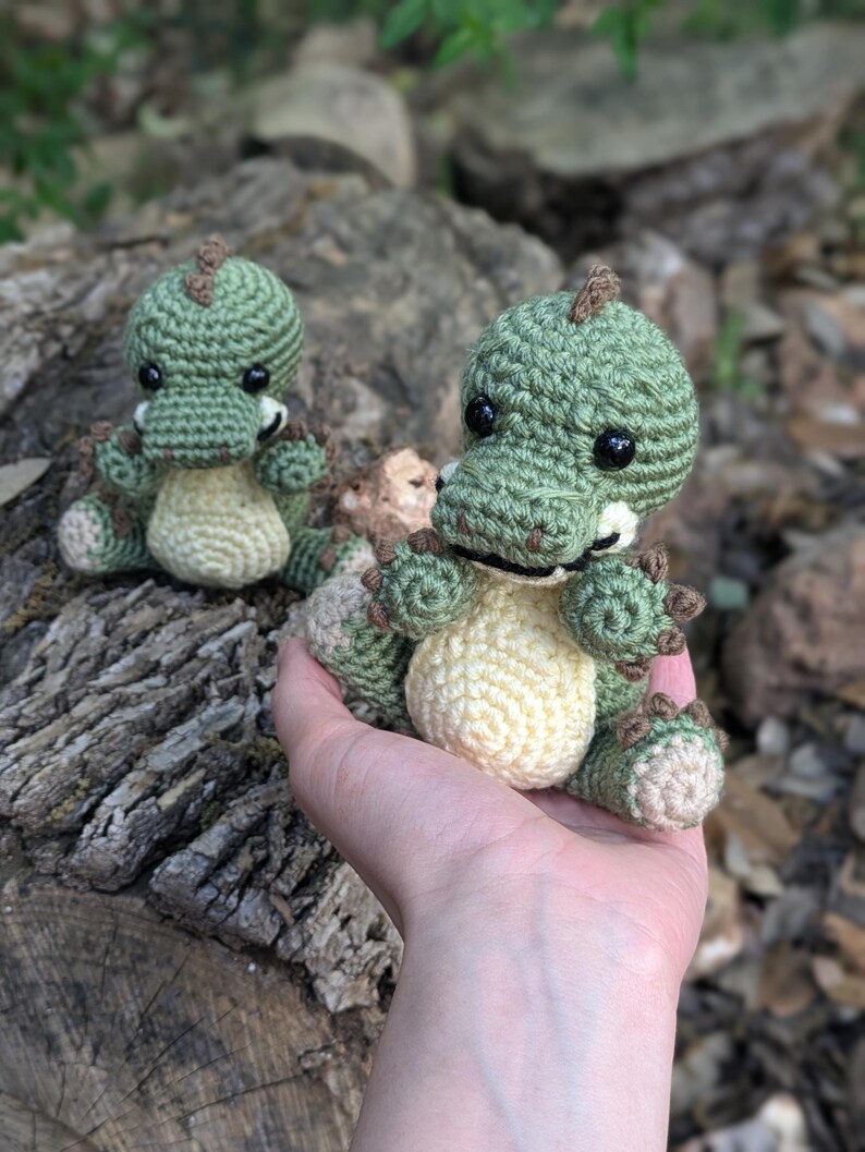 May include: Two crocheted green dinosaur plushies with brown accents. The smaller dinosaur is sitting on a tree stump, while the larger dinosaur is being held in a hand.