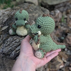 May include: Two crocheted green dinosaur toys. The larger dinosaur is being held in a hand. Both dinosaurs have brown accents and black eyes.