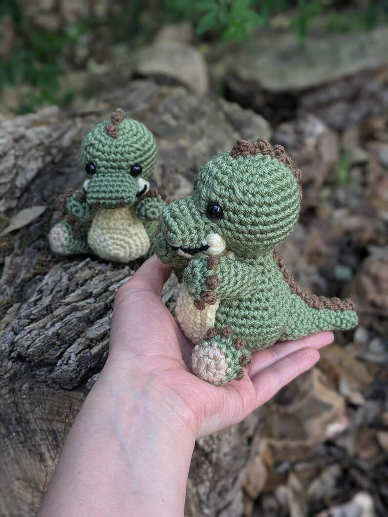 May include: Two crocheted green dinosaur toys. The larger dinosaur is being held in a hand. Both dinosaurs have brown accents and black eyes.
