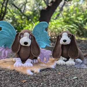 May include: Two crochet basset hound dog stuffed animals. One has blue wings and purple accents. Both dogs are brown, white, and black. They are sitting on a faux fur rug. The background is a blurred green forest.