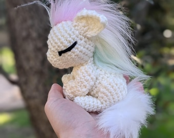May include: A small, cream-colored crocheted unicorn with a pink and pastel-colored mane and tail. The unicorn has closed black eyes and is held in a hand against a blurred green background. The plush toy is a handmade craft.