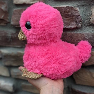 May include: A pink plush duck with gold glitter accents. The duck has large black eyes and a fluffy texture.