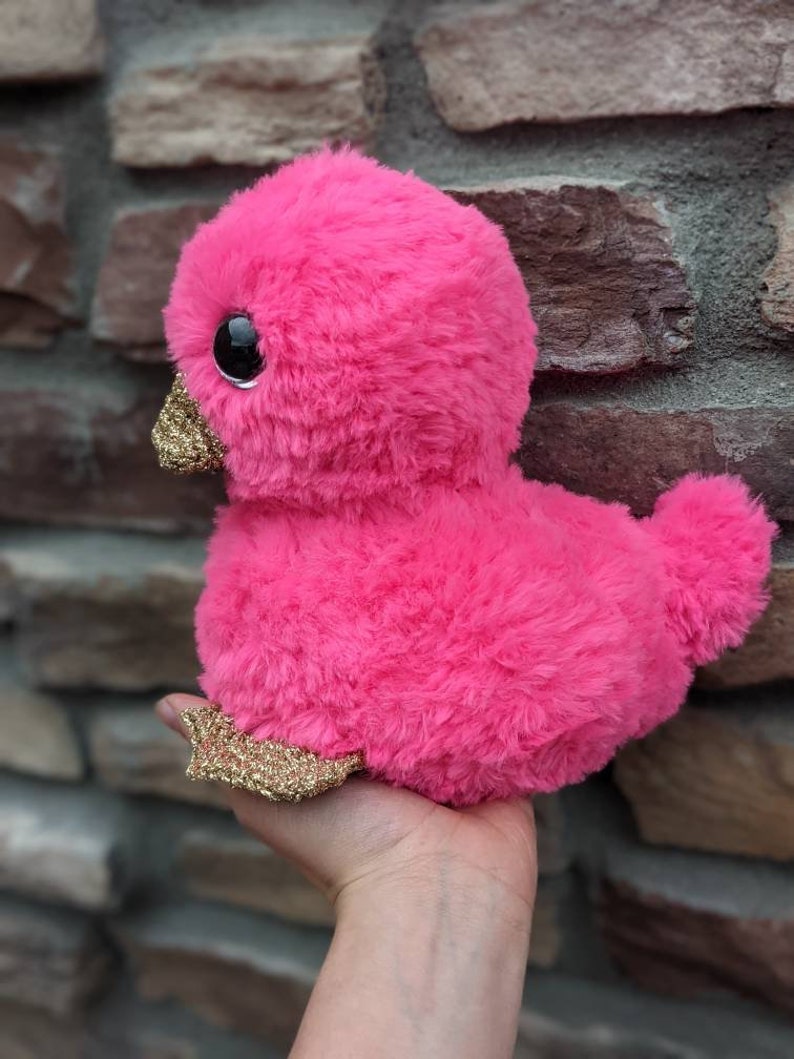 May include: A pink plush duck with gold glitter accents. The duck has large black eyes and a fluffy texture.