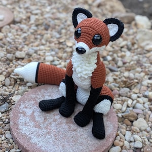 May include: A crocheted fox figurine featuring a rust-orange body, a white chest with textured stitches, black legs, and black-tipped ears. The fox has large, glossy black safety eyes and a black nose, with a long tail ending in a white tip.