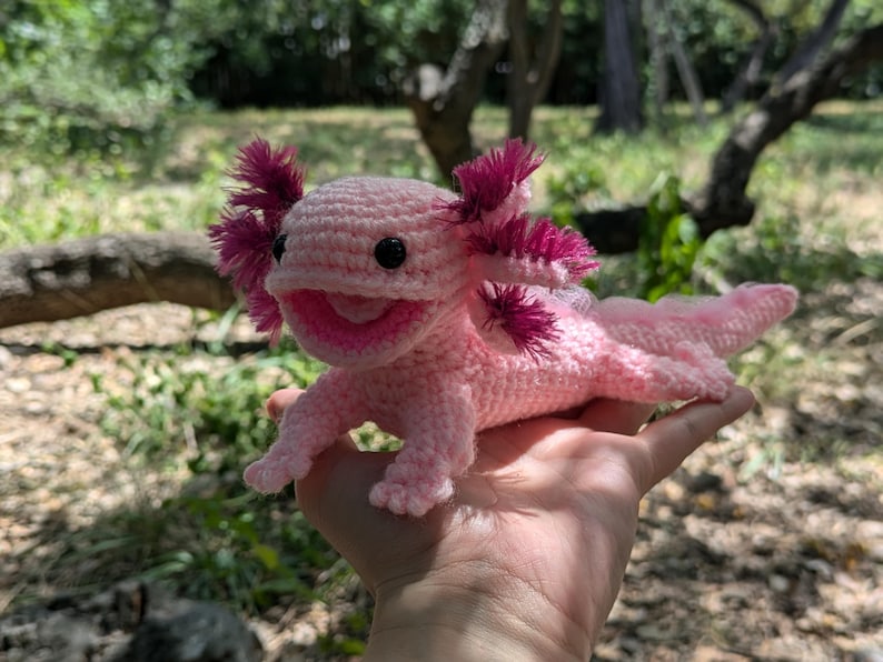 Może przedstawiać: R&oacute;żowa pluszowa zabawka axolotl wykonana na szydełku z r&oacute;żowymi i fioletowymi akcentami. Zabawka ma uśmiechniętą twarz i jest trzymana w dłoni osoby.