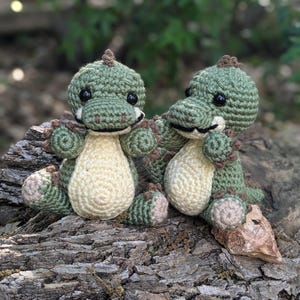 May include: Two green crocheted dinosaur toys with black eyes and brown noses sitting on a tree trunk.