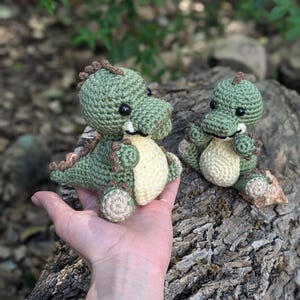 May include: Two crocheted green dinosaur toys with brown accents. The larger dinosaur is being held in a hand, while the smaller dinosaur is sitting on a tree trunk.