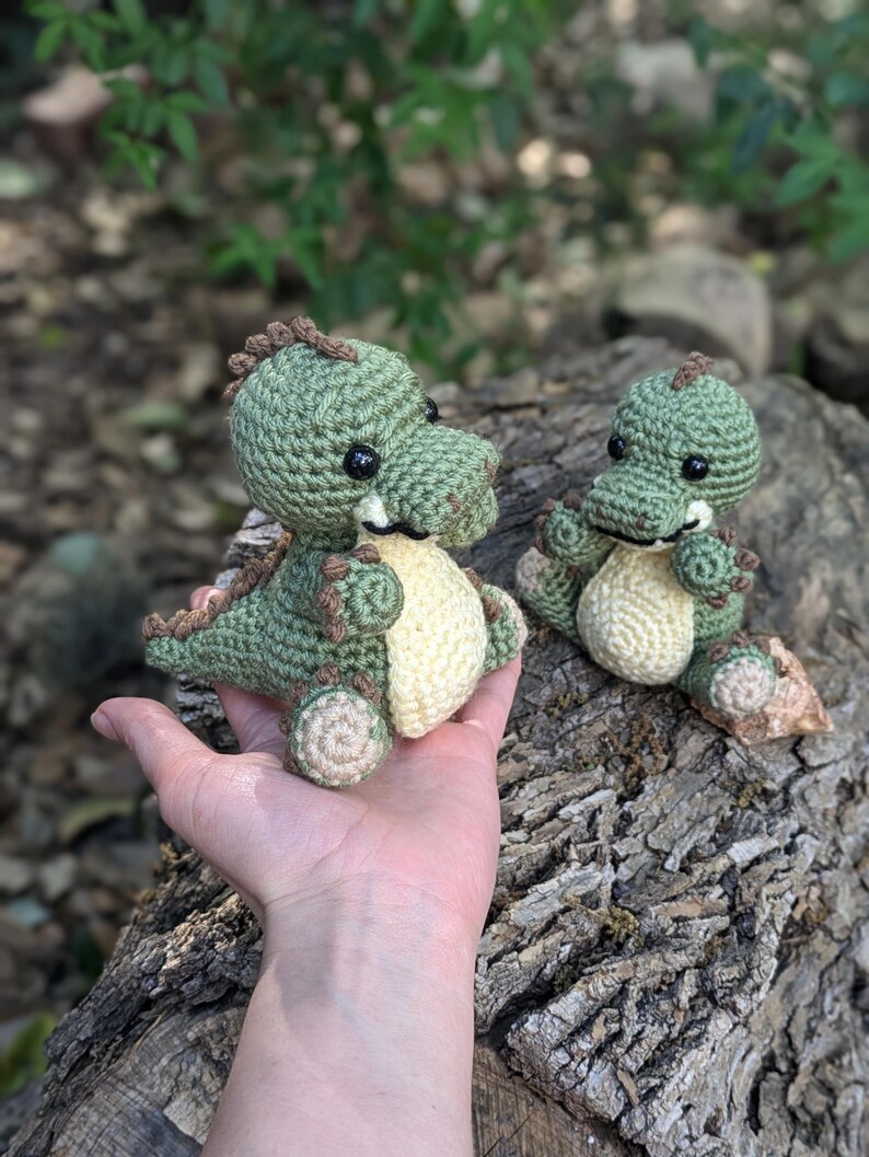 May include: Two crocheted green dinosaur toys with brown accents. The larger dinosaur is being held in a hand, while the smaller dinosaur is sitting on a tree trunk.