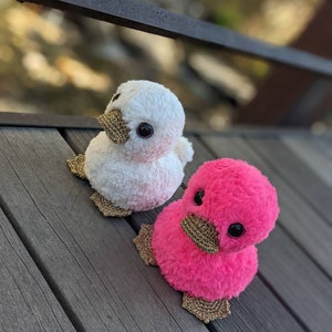 May include: Two plush duck toys, one white and one pink, with gold glitter accents. The ducks are sitting on a wooden surface.