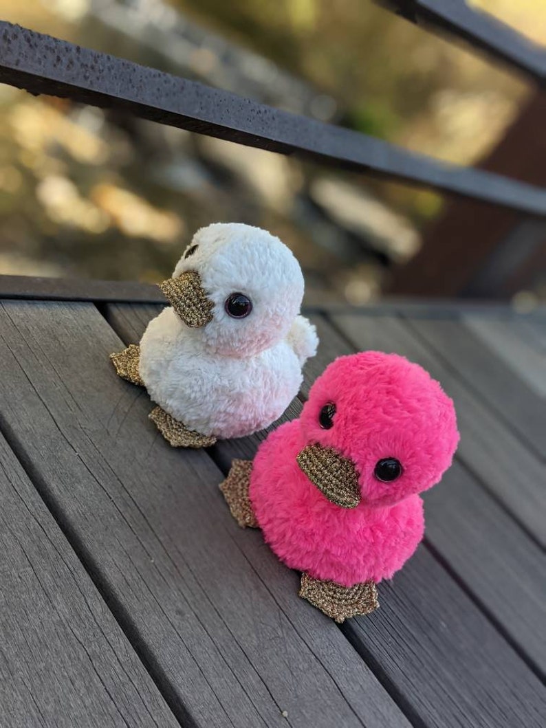 May include: Two plush duck toys, one white and one pink, with gold glitter accents. The ducks are sitting on a wooden surface.
