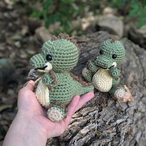 May include: Two crocheted green dinosaur toys with brown accents. The larger dinosaur is being held in a hand, while the smaller dinosaur is sitting on a tree stump.