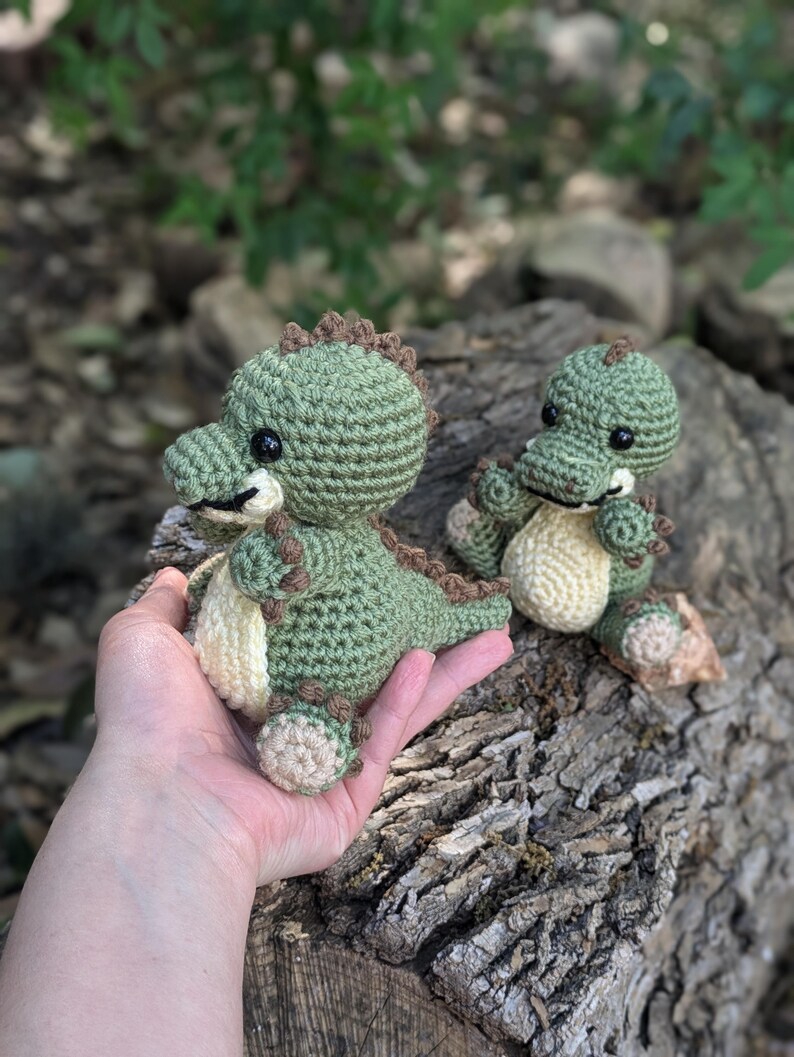 May include: Two crocheted green dinosaur toys with brown accents. The larger dinosaur is being held in a hand, while the smaller dinosaur is sitting on a tree stump.