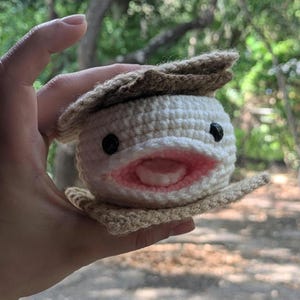 May include: A crocheted axolotl toy, white with a pink mouth and black eyes, held in a hand. It features a tan frill. This handmade plushie is a unique, handcrafted item.