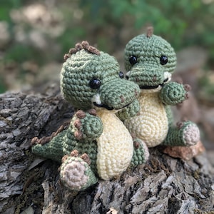 May include: Two small green crocheted dinosaur toys with black eyes and brown noses sitting on a tree stump.