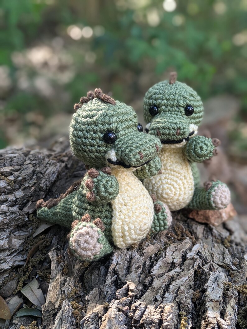 May include: Two small green crocheted dinosaur toys with black eyes and brown noses sitting on a tree stump.