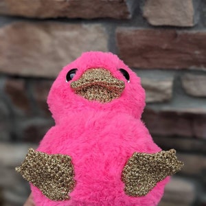May include: A pink plush toy shaped like a duck with gold glitter accents on its wings and beak.