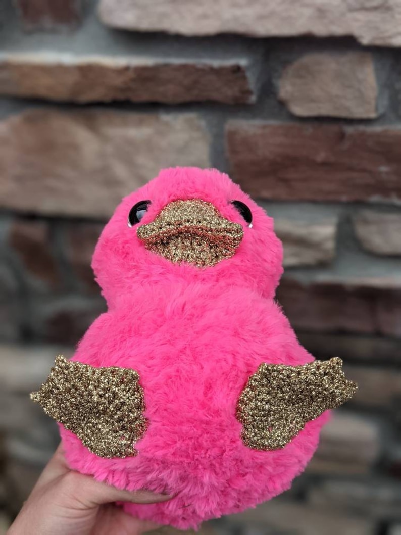 May include: A pink plush toy shaped like a duck with gold glitter accents on its wings and beak.