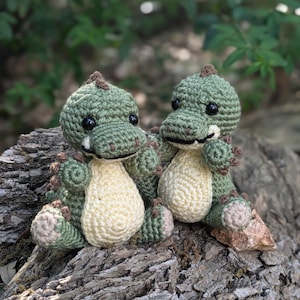 May include: Two small green crocheted dinosaur toys sitting on a tree trunk. The dinosaurs have brown eyes and brown accents on their feet and tails.