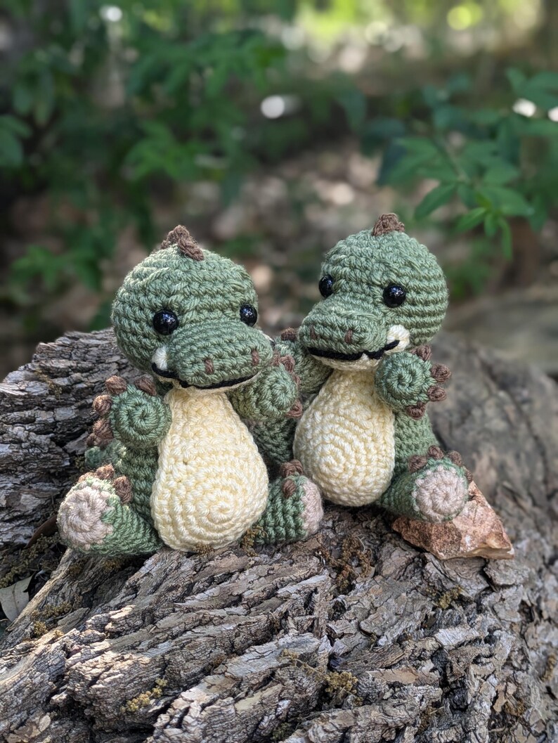 May include: Two small green crocheted dinosaur toys sitting on a tree trunk. The dinosaurs have brown eyes and brown accents on their feet and tails.