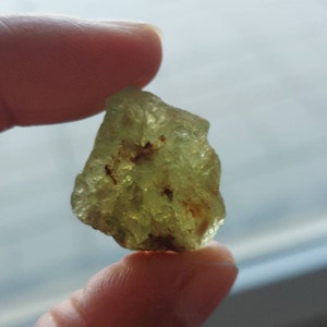 Pode incluir: Uma gema verde translúcida bruta com inclusões marrons. A pedra tem uma forma irregular e uma aparência ligeiramente nublada.