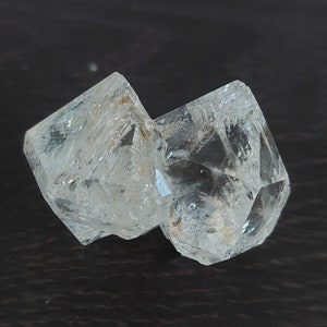 Może przedstawiać: Dwa przezroczyste, fasetowane kryształy, jeden nieco większy od drugiego, są połączone ze sobą. Kryształy mają lekko zamglony wygląd, z odcieniem niebieskiego.