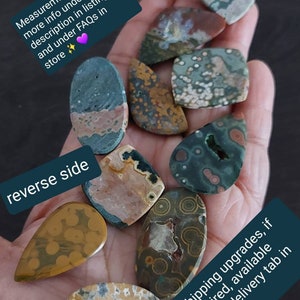 May include: A collection of eight green, brown, and white ocean jasper cabochons with various patterns. The cabochons are displayed on a hand.