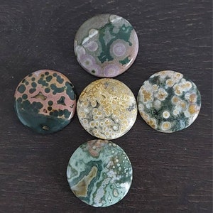 May include: Five round, polished ocean jasper stones with various patterns of green, brown, pink, and white. The stones are arranged in a cross shape on a dark brown surface.