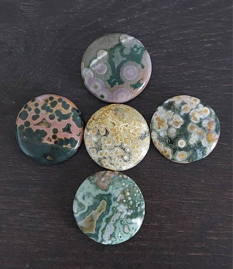 May include: Five round, polished ocean jasper stones with various patterns of green, brown, pink, and white. The stones are arranged in a cross shape on a dark brown surface.
