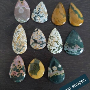 May include: Twelve pear-shaped ocean jasper stones in various shades of green, brown, and white with a speckled pattern. The stones are arranged in three rows of four on a dark brown surface. The text "Pear shapes" is written in white on a black background at the bottom of the image.